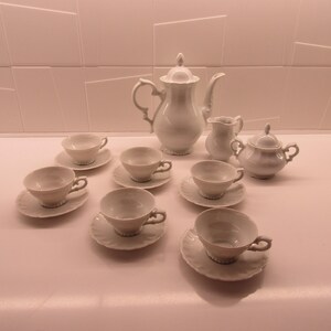 May include: A white porcelain tea set consisting of a tall teapot with a lid, a creamer, a lidded sugar bowl, and six teacups with matching saucers. Each piece features a smooth, glossy finish and delicate, curved handles on the cups and pots.