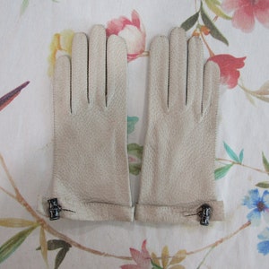 May include: A pair of light beige leather gloves with a textured surface. The gloves have a cuff with a small decorative clasp. The gloves are displayed on a floral patterned fabric.