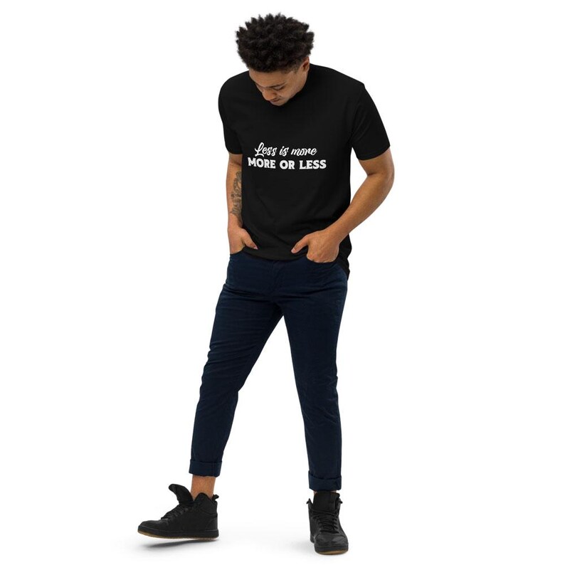 K&ouml;nnte beinhalten: Ein schwarzes T-Shirt mit dem wei&szlig;en Text "Less is more MORE OR LESS". Das Shirt wird mit dunkelblauen Hosen und schwarzen Schuhen kombiniert. Die Person steht mit den H&auml;nden in den Taschen.