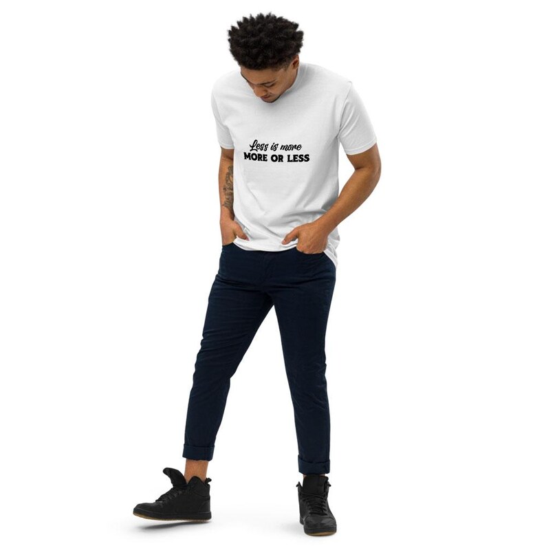 K&ouml;nnte beinhalten: Wei&szlig;es T-Shirt mit dem Text "Less is more MORE OR LESS" in schwarzer Schrift. Die Person tr&auml;gt eine marineblaue Hose und schwarze Schuhe. Das T-Shirt ist ein l&auml;ssiger Stil.
