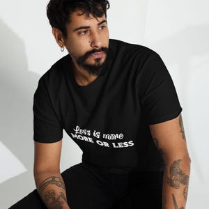 K&ouml;nnte beinhalten: Schwarzes T-Shirt mit dem wei&szlig;en Text "Less is more MORE OR LESS". Das Shirt hat einen Rundhalsausschnitt und scheint aus einem weichen Material zu sein. Das Bild zeigt das Shirt an einer Person.