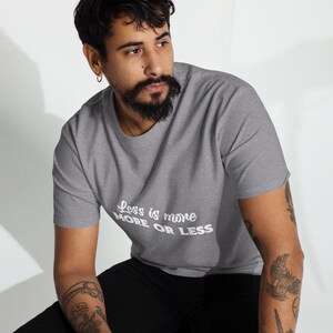K&ouml;nnte beinhalten: Graues T-Shirt mit dem wei&szlig;en Schriftzug "Less is more MORE OR LESS". Das kurz&auml;rmelige Shirt scheint aus einem weichen Material zu sein. Die Person, die das Shirt tr&auml;gt, sitzt.