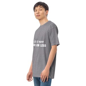 K&ouml;nnte beinhalten: Ein grau meliertes T-Shirt mit dem wei&szlig;en Text "Less is more, MORE OR LESS". Das T-Shirt hat kurze &Auml;rmel und einen Rundhalsausschnitt. Die Person tr&auml;gt eine blaue Jeans.