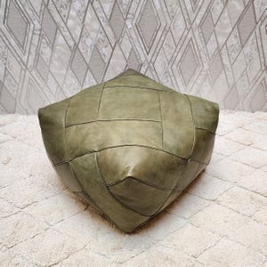 Pode incluir: Um pufe quadrado de couro verde-oliva com um design de patchwork geométrico. O pufe está colocado sobre um tapete branco texturizado. O fundo apresenta um papel de parede estampado.