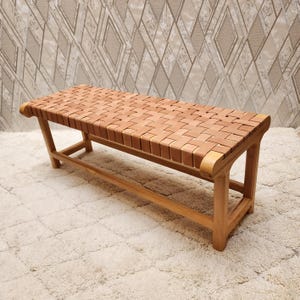 Puede incluir: Un banco marrón claro con un asiento de cuero tejido y un marco de madera. El banco tiene forma rectangular con bordes curvos en los extremos. Las patas están en ángulo y conectadas por un travesaño inferior.