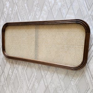 May include: Rectangular wall decor featuring a woven cane center framed by a dark brown wooden border. The cane has a light beige color, and the frame has rounded corners. The piece is mounted on a patterned wallpaper background.