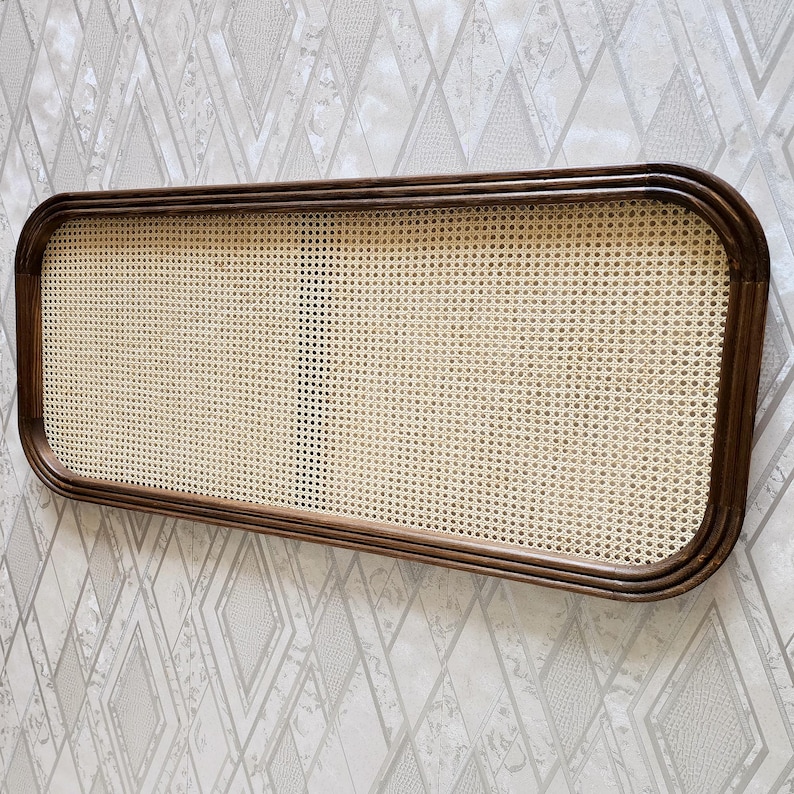 May include: Rectangular wall decor featuring a woven cane center framed by a dark brown wooden border. The cane has a light beige color, and the frame has rounded corners. The piece is mounted on a patterned wallpaper background.