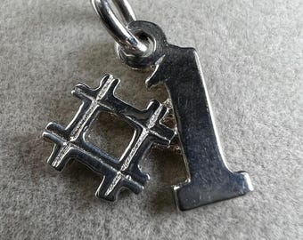 May include: A silver charm featuring the number one and a hashtag symbol. The charm has a loop at the top for attaching to a necklace or bracelet. The metal has a shiny finish.