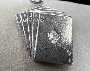 May include: A silver-toned pendant featuring a fan of playing cards. The cards are marked with the Jack, Jack, King, and Ace of Spades. The pendant has a small loop at the top for a chain.