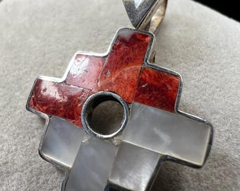 May include: A silver pendant with a cross-shaped design. The cross features red and white inlays, with a circular hole in the center. The pendant has a teardrop-shaped bail at the top for a chain.