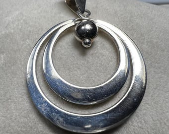 May include: A silver pendant featuring two nested, polished rings. A small, round bead sits between the rings, and a loop for a chain is at the top. The pendant has a simple, modern design.
