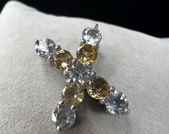May include: A silver-toned pendant in the shape of the letter "X", adorned with alternating light blue and yellow gemstones. The gemstones are round-cut and set closely together, creating a sparkling effect. The pendant is displayed on a light gray surface.