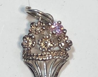 May include: A silver-toned charm in the shape of a flower basket. The basket is filled with detailed flowers, including one prominent pink gemstone. The charm has a loop for attaching to a bracelet or necklace.