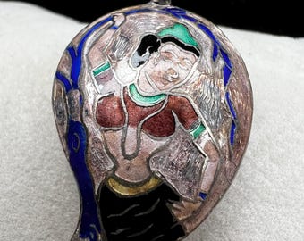 May include: A silver-toned pendant with a detailed enamel design. The pendant features a dancing figure in red, black, and green attire, set against a silver background with blue accents. The pendant has a loop at the top for hanging.