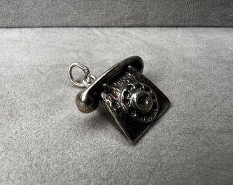 May include: A silver-toned charm in the shape of a vintage rotary phone. The charm features a detailed dial, receiver, and a loop for attaching to a necklace or bracelet. The phone is a classic design.