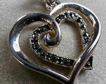 May include: A silver heart-shaped pendant with a swirling design. The pendant features a double heart outline, with the inner heart embellished with small, dark gemstones. The bail is attached at the top for a necklace chain.
