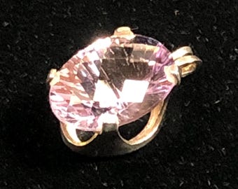 May include: A light pink gemstone ring with a gold-colored band. The gemstone is oval-shaped and faceted, reflecting light. The ring is set against a dark background, highlighting its sparkle and design.