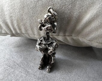 May include: A silver-colored pendant in the shape of a seated figure with a textured, abstract design. The figure has a loop at the top for a chain and is resting on a gray surface.