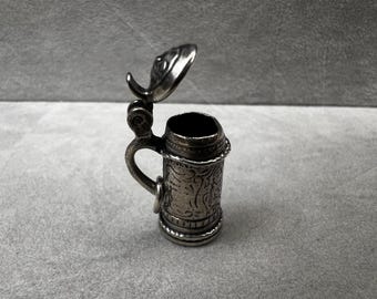 May include: A small, silver-colored miniature beer stein with an open lid. The stein has a handle and is decorated with an embossed floral pattern. The lid is shaped like a leaf. The stein is on a gray surface.