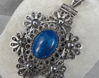 May include: Silver-toned pendant with a large, oval, blue gemstone center. The pendant features a filigree design with floral accents and small, round embellishments. The bail is intricately detailed. The pendant is displayed on a soft, light-colored surface.