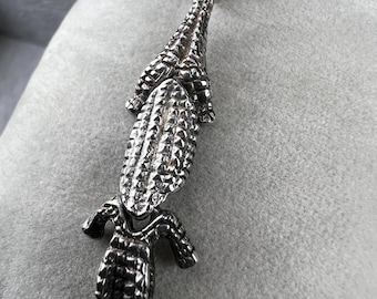 May include: A silver-colored pendant in the shape of an alligator. The pendant has a textured surface, with detailed scales and limbs. It is suspended from a small, silver-colored loop.