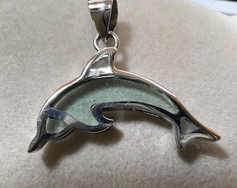May include: A silver dolphin pendant with a light green center. The pendant is suspended from a silver bail. The dolphin's body is outlined in silver, with a smooth, polished finish. The pendant is displayed on a light gray surface.
