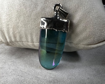 May include: A pendant with a polished, iridescent blue-green glass body and a silver-colored metal cap and bail. The glass has a smooth, rounded shape. The pendant is displayed on a light gray surface.