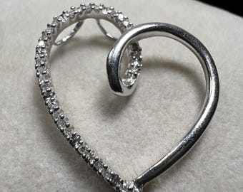 May include: A silver heart-shaped pendant. One side of the heart is encrusted with small, clear gemstones, while the other side is a smooth, polished silver. The pendant is displayed on a soft, light-colored surface.