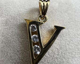 May include: Gold-toned pendant in the shape of the letter "V", featuring three clear, round gemstones. The pendant has a polished finish and a decorative bail for a chain. The background is a soft, neutral color.