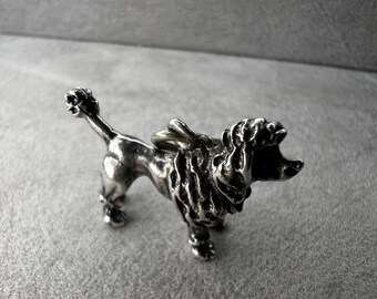May include: A silver-colored poodle figurine. The dog is depicted in a standing pose, with its head turned slightly to the side. The poodle has a detailed, textured coat and is a decorative item.