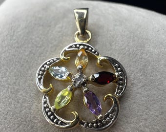 May include: A gold and silver pendant with a flower design. The pendant features a central clear stone surrounded by six colorful gemstones: yellow, green, purple, red, blue, and orange. The pendant is on a neutral background.