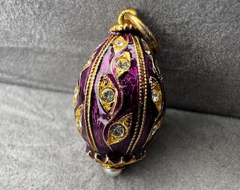 May include: A purple and gold pendant with a teardrop shape. The pendant is adorned with gold accents and clear, sparkling gemstones. A small, white pearl hangs from the bottom. The pendant has a gold loop at the top for hanging.