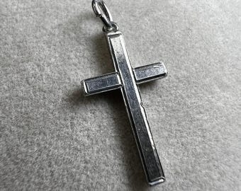May include: A silver-colored cross pendant with a loop at the top for a chain. The cross has a simple, classic design with a rectangular shape and a polished finish. The pendant is displayed on a light gray surface.