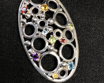May include: Silver-toned pendant with an oval shape, featuring a series of interconnected circles and spheres. The pendant is adorned with various colorful gemstones, including red, yellow, blue, green, and purple. The pendant has a loop at the top for a chain.