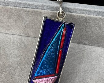 May include: A silver pendant with a rectangular, dark blue enamel background. The pendant features a sailboat design with a blue and pink sail and a red mast. The pendant has a silver bail for hanging.