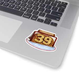 May include: A sticker featuring a hockey net with the text "Lumberyard 39." The number 39 is made of a textured, light brown material, resembling wood. The net is red and white, with a blue and white base.