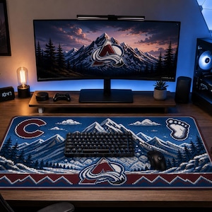 May include: A computer desk setup featuring a large blue and red Colorado Avalanche mousepad with a mountain design, a black keyboard, and a computer monitor displaying a mountain scene. The desk also includes a computer tower, speakers, and a mug.