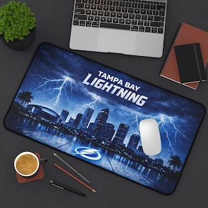 May include: A rectangular Tampa Bay Lightning mousepad featuring a cityscape under a stormy sky with lightning bolts. The team's name is displayed in white. A computer mouse rests on the pad, alongside a laptop, coffee, and stationery.