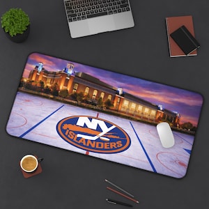 May include: A rectangular mouse pad featuring a hockey rink design with the NY Islanders logo. The background shows a building at dusk with a colorful sky. A computer mouse and keyboard are also visible.