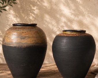 May include: Two ceramic vases with a rustic design. The upper portion of the vases is a mix of brown and tan, while the lower half is black. The vases have a rounded shape and a dark rim. They are placed on a wooden surface.