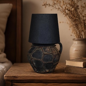May include: A table lamp featuring a dark, textured, spherical ceramic base with a grid-like pattern of raised lines and a single handle. The lamp is topped with a simple, solid black, tapered cylindrical lampshade that provides a focused light.