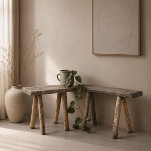May include: A weathered wooden bench with three legs, alongside a smaller bench, and a green ceramic mug. A large beige vase with dried branches and a framed abstract artwork complete the neutral interior.