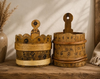 Puede incluir: Dos cubos decorativos de madera con patrones florales y geométricos. El cubo de la izquierda tiene una parte superior festoneada y un asa decorativa. El cubo de la derecha tiene un asa y está decorado con diseños florales. Ambos están sobre una superficie de madera.