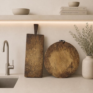 Puede incluir: Dos tablas de cortar de madera desgastada, una rectangular con mango y otra circular con un lazo de cuerda negra, se apoyan sobre una encimera clara junto a un grifo de cocina y un jarrón de cerámica con ramas verdes.