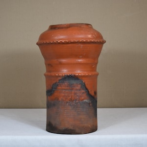May include: A terracotta-coloured, cylindrical vase with a textured surface. The vase has a decorative beaded rim and a dark, mottled area near the base. The vase is displayed on a white surface.