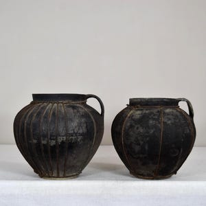 Könnte beinhalten: Zwei antike schwarze Töpferwarenvasen mit Griffen. Die Vasen haben eine abgerundete Form mit vertikalen Rippendetails. Die Vase links hat eine ausgeprägtere Form und ist etwas größer als die Vase rechts.
