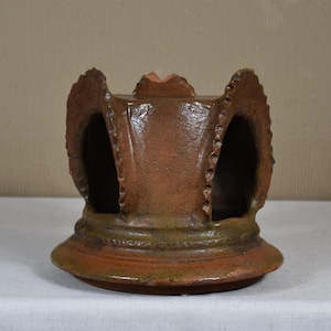 Puede incluir: Un portavelas de cerámica marrón hecho a mano con un diseño abierto único. El portavelas presenta una base ancha y circular y una serie de aberturas arqueadas en los lados. La parte superior tiene un borde decorativo y texturizado.