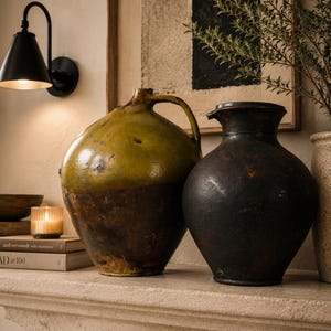 May include: Two antique ceramic vases on a stone shelf. One is a large, round jug with a handle, glazed in olive green and brown. The other is a dark, matte black vase with a narrow neck. A lit candle and books are in the background.