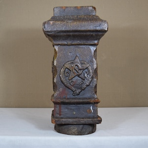 May include: A weathered, dark brown, rectangular pillar with a decorative emblem featuring a star and serpent. The pillar has a textured surface and is set on a white surface.