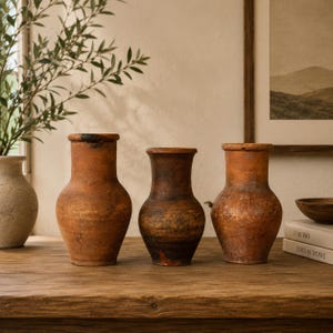 May include: Three rustic terracotta vases with a weathered appearance. The vases are various shades of brown and have a classic, slightly flared shape. They sit on a wooden surface, suggesting a vintage or farmhouse aesthetic.